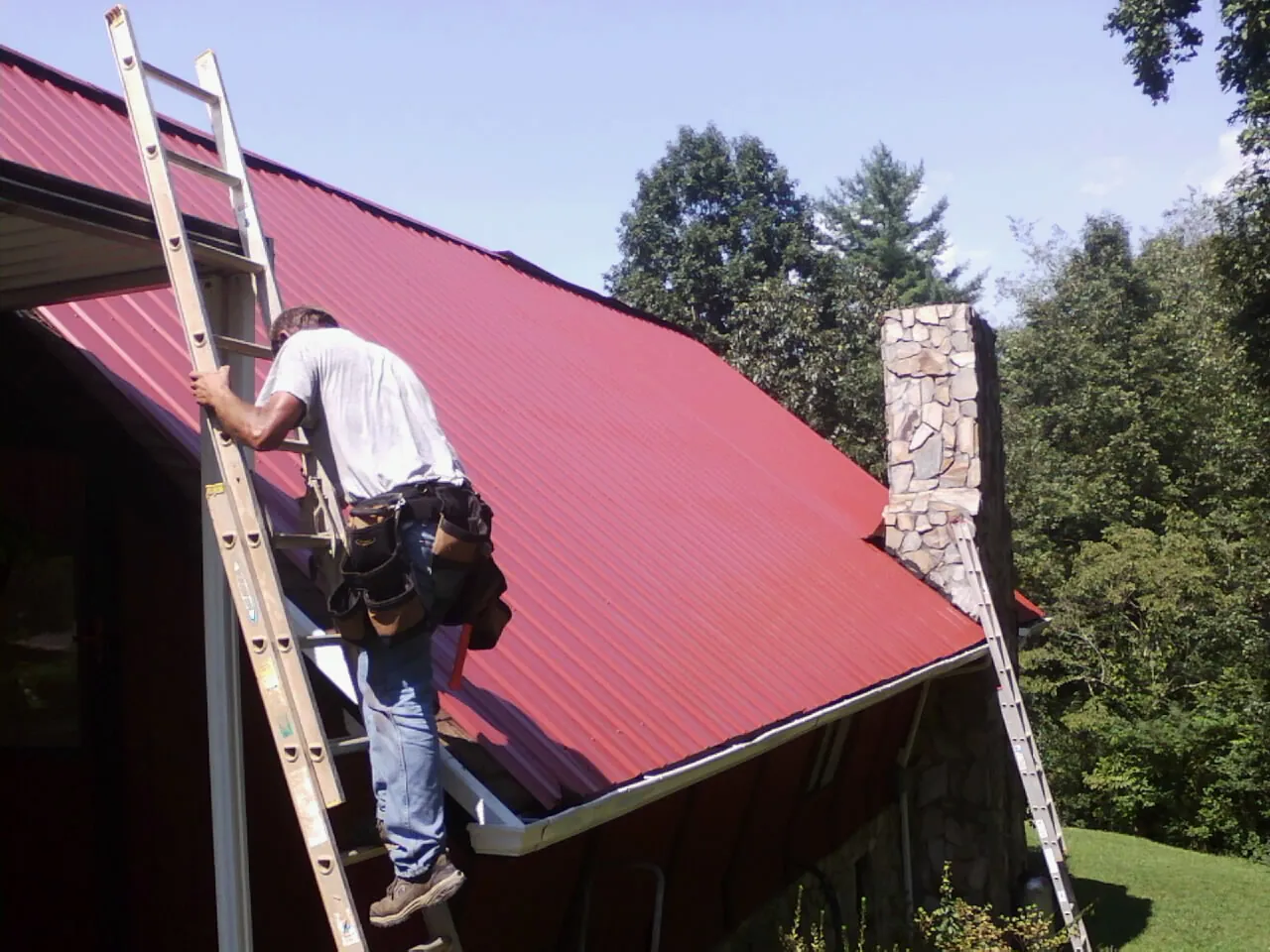 Roofer on a ladder beside a red metal roof and stone chimney — Soffit & Fascia Repair by Roofing Brothers in New Stuyahok